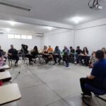 A imagem mostra o professor Márcio Fábio Belos Matos durante entrevista para o e-book sobre os 20 anos do curso de Jornalismo da UFMA. Ao redor dele, alunos estão sentados em círculo acompanhando a conversa, enquanto uma câmera grava a entrevista.