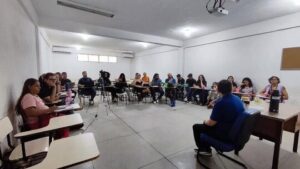 A imagem mostra o professor Márcio Fábio Belos Matos durante entrevista para o e-book sobre os 20 anos do curso de Jornalismo da UFMA. Ao redor dele, alunos estão sentados em círculo acompanhando a conversa, enquanto uma câmera grava a entrevista.