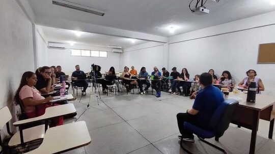 A imagem mostra o professor Márcio Fábio Belos Matos durante entrevista para o e-book sobre os 20 anos do curso de Jornalismo da UFMA. Ao redor dele, alunos estão sentados em círculo acompanhando a conversa, enquanto uma câmera grava a entrevista.